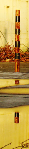 A yellow and black striped metal post erected in the concrete next to an abandoned warehouse. Pools of water nearby reflect the post's image. We had to sneak behind some chainlink fences for this picture.
