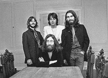 A photo of John Lennon seated at a table with the rest of the Beatles standing around him.