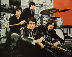 A picture of the members of the Kinks resting on a stage around a drumset with their instruments.