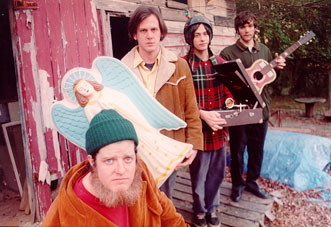 Picture of band members from Neutral Milk Hotel standing around a rundown shack holding various items.